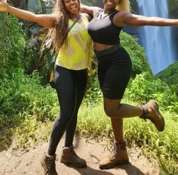Two female tourists posing joyfully with arms outstretched in front of Sipi Falls waterfall in eastern Uganda during a guided hiking tour